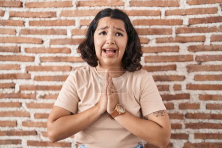 Photo for Young hispanic woman standing over bricks wall begging and praying with hands together with hope expression on face very emotional and worried. begging. - Royalty Free Image