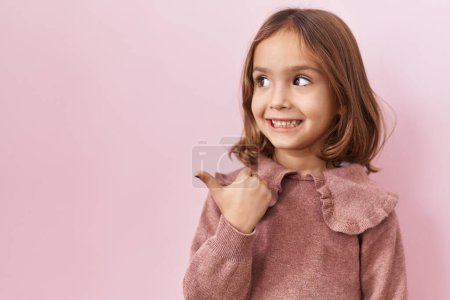 Photo for Little hispanic girl standing over pink background smiling with happy face looking and pointing to the side with thumb up. - Royalty Free Image