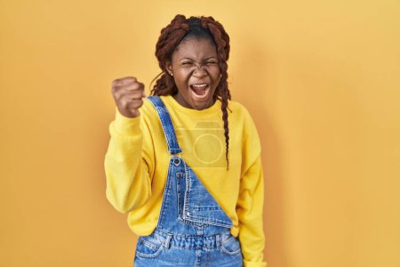 Foto de Mujer africana de pie sobre fondo amarillo enojado y loco levantando el puño frustrado y furioso mientras gritaba con ira. rabia y concepto agresivo. - Imagen libre de derechos