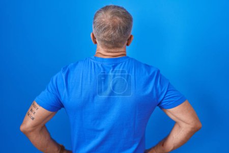 Téléchargez les photos : Homme hispanique avec les cheveux gris debout sur fond bleu debout vers l'arrière regardant loin avec les bras sur le corps - en image libre de droit