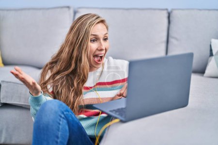 Téléchargez les photos : Jeune femme blonde utilisant un ordinateur portable à la maison célébrant la réalisation avec un sourire heureux et l'expression gagnante avec la main levée - en image libre de droit