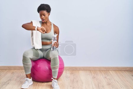 Foto de Mujer afroamericana usando ropa deportiva sentada en la pelota de pilates mirando el tiempo del reloj preocupado, con miedo de llegar tarde - Imagen libre de derechos