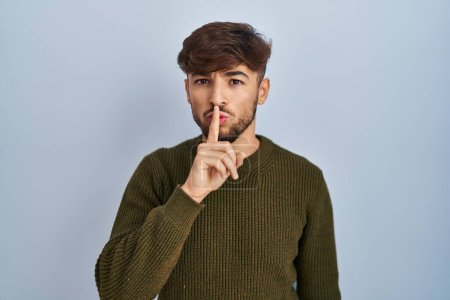 Téléchargez les photos : Homme arabe avec la barbe debout sur fond bleu demandant à être calme avec le doigt sur les lèvres. silence et concept secret. - en image libre de droit