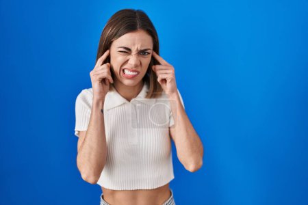 Foto de Hispanic woman standing over blue background covering ears with fingers with annoyed expression for the noise of loud music. deaf concept. - Imagen libre de derechos