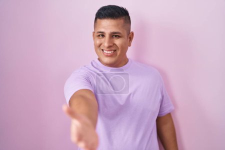 Téléchargez les photos : Young hispanic man standing over pink background smiling cheerful offering palm hand giving assistance and acceptance. - en image libre de droit