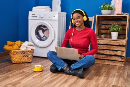 Photo for African american woman using laptop waiting for washing machine at laundry room - Royalty Free Image