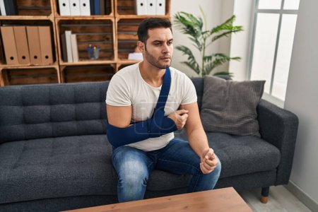 Photo for Young hispanic man with beard wearing arm on sling sitting at therapy consult thinking attitude and sober expression looking self confident - Royalty Free Image