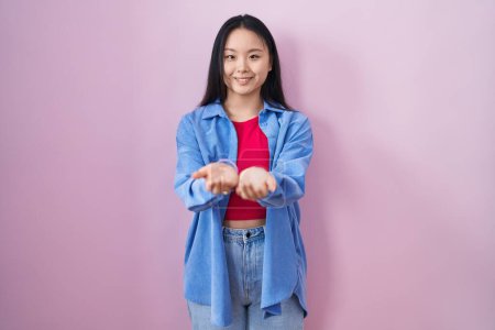 Téléchargez les photos : Jeune femme asiatique debout sur fond rose souriant avec les mains paumes ensemble recevant ou donnant geste. maintien et protection - en image libre de droit