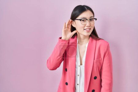 Téléchargez les photos : Jeune femme d'affaires chinoise portant des lunettes souriant avec la main sur l'oreille en écoutant une rumeur ou des commérages. concept de surdité. - en image libre de droit