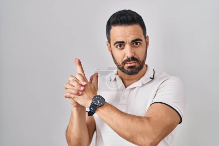 Téléchargez les photos : Jeune homme hispanique avec barbe portant des vêtements décontractés sur fond blanc tenant un pistolet symbolique avec geste de la main, jouant à tuer des armes de tir, visage en colère - en image libre de droit