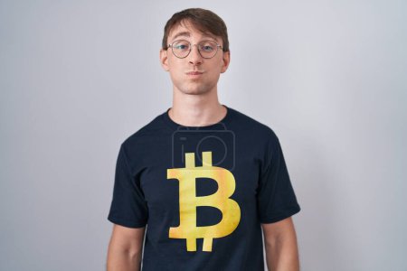 Téléchargez les photos : Homme blond caucasien portant Bitcoin t-shirt gonflant les joues avec un visage drôle. bouche gonflée d'air, expression folle. - en image libre de droit