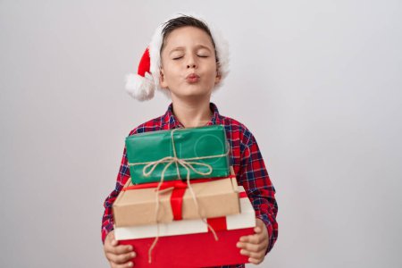Téléchargez les photos : Petit garçon hispanique portant un chapeau de Noël tenant des cadeaux en regardant la caméra souffler un baiser étant charmant et sexy. expression d'amour. - en image libre de droit