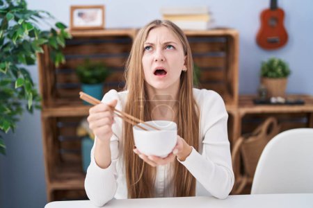 Foto de Joven mujer caucásica comiendo comida asiática usando palillos enojado y loco gritando frustrado y furioso, gritando con ira mirando hacia arriba. - Imagen libre de derechos