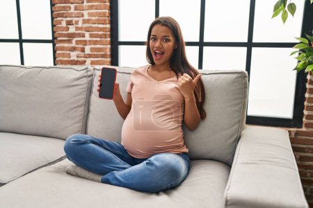 Foto de Mujer embarazada joven sosteniendo teléfono inteligente que muestra la pantalla apuntando pulgar hacia el lado sonriendo feliz con la boca abierta - Imagen libre de derechos