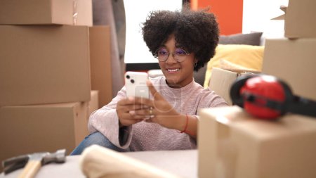 Foto de Mujer afroamericana joven sonriendo confiada usando teléfono inteligente en su nuevo hogar - Imagen libre de derechos