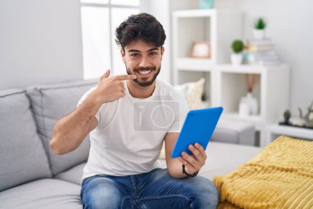 Photo for Hispanic man with beard using touchpad sitting on the sofa smiling cheerful showing and pointing with fingers teeth and mouth. dental health concept. - Royalty Free Image