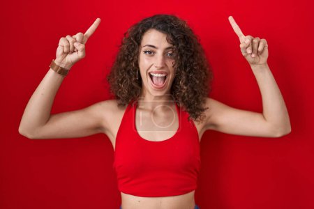Foto de Mujer hispana con el pelo rizado de pie sobre fondo rojo sonriendo asombrada y sorprendida y señalando con los dedos y los brazos levantados. - Imagen libre de derechos