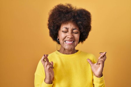 Téléchargez les photos : Jeune femme afro-américaine debout sur fond jaune geste doigt croisé souriant avec espoir et les yeux fermés. chance et concept superstitieux. - en image libre de droit