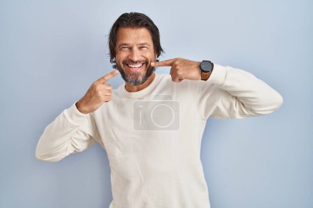 Téléchargez les photos : Bel homme d'âge moyen portant pull décontracté sur fond bleu souriant gai montrant et pointant avec les doigts dents et la bouche. concept de santé dentaire. - en image libre de droit