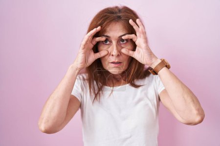 Foto de Middle age woman standing over pink background trying to open eyes with fingers, sleepy and tired for morning fatigue - Imagen libre de derechos