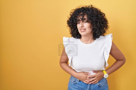 Téléchargez les photos : Jeune femme du Moyen-Orient debout sur fond jaune avec la main sur le ventre parce que l'indigestion, la maladie douloureuse se sent mal. concept de douleur. - en image libre de droit