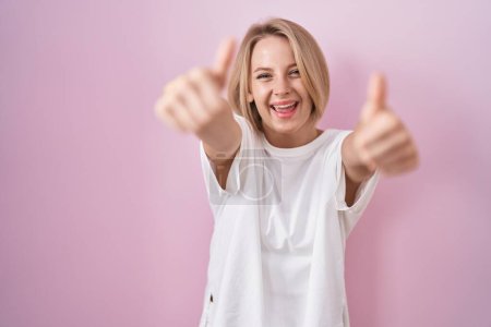 Photo for Young caucasian woman standing over pink background approving doing positive gesture with hand, thumbs up smiling and happy for success. winner gesture. - Royalty Free Image