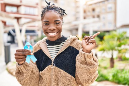 Foto de Hermosa mujer negra sosteniendo la cinta azul sonriendo feliz señalando con la mano y el dedo hacia un lado - Imagen libre de derechos