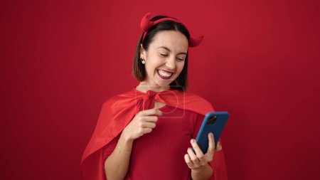 Téléchargez les photos : Jeune belle femme hispanique portant costume diable en utilisant smartphone sur fond rouge isolé - en image libre de droit
