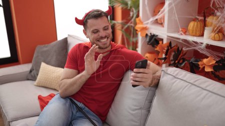 Foto de Joven caucásico vestido de diablo sentado en el sofá haciendo videollamada en casa - Imagen libre de derechos