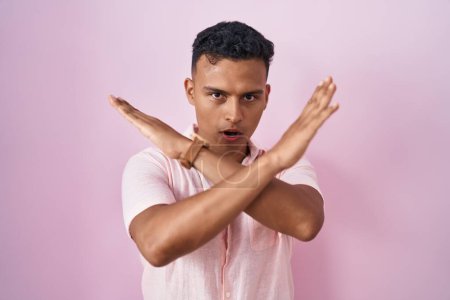 Téléchargez les photos : Young hispanic man standing over pink background rejection expression crossing arms doing negative sign, angry face - en image libre de droit