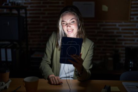 Foto de Mujer rubia caucásica que trabaja en la oficina por la noche sacando la lengua feliz con expresión divertida. concepto de emoción. - Imagen libre de derechos