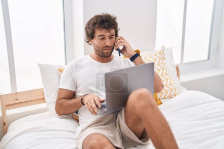 Photo for Young hispanic man using laptop talking on smartphone at bedroom - Royalty Free Image