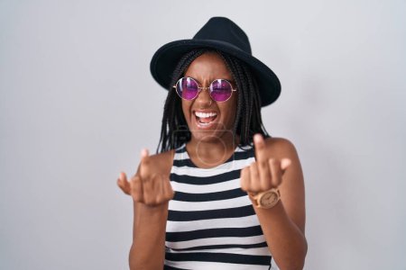 Foto de Young african american with braids wearing hat and sunglasses showing middle finger doing fuck you bad expression, provocation and rude attitude. screaming excited - Imagen libre de derechos