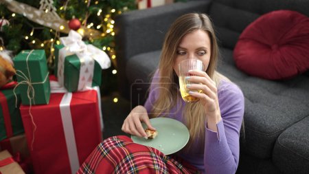 Téléchargez les photos : Jeune femme blonde prenant le petit déjeuner assis sur le sol près de l'arbre de Noël à la maison - en image libre de droit
