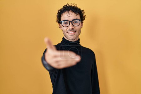 Foto de Hombre hispano de pie sobre fondo amarillo sonriente amistoso ofreciendo apretón de manos como saludo y bienvenida. negocios exitosos. - Imagen libre de derechos