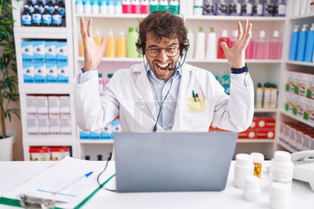 Téléchargez les photos : Jeune homme hispanique travaillant à la pharmacie pharmacie travaillant avec ordinateur portable célébrant la victoire avec sourire heureux et expression gagnante avec les mains levées - en image libre de droit