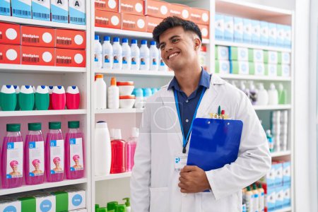 Foto de Joven farmacéutico hispano sonriendo confiado sosteniendo portapapeles en la farmacia - Imagen libre de derechos