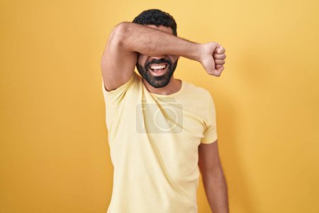 Téléchargez les photos : Homme hispanique avec barbe debout sur fond jaune couvrant les yeux avec le bras souriant gai et drôle. concept aveugle. - en image libre de droit