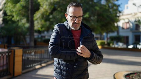 Foto de Hombre de mediana edad de pie con expresión seria buscando algo en la chaqueta en el parque - Imagen libre de derechos
