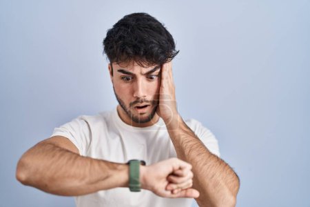 Foto de Hombre hispano con barba de pie sobre fondo blanco mirando el tiempo del reloj preocupado, con miedo de llegar tarde - Imagen libre de derechos