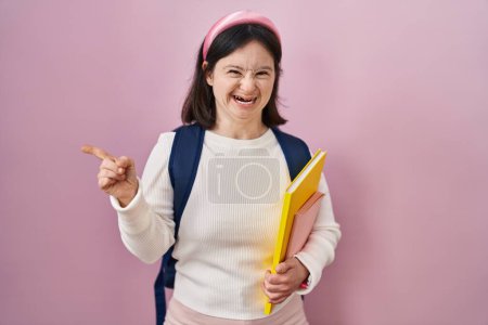 Foto de Mujer con síndrome de Down usando mochila de estudiante y sosteniendo libros señalando a un lado preocupado y nervioso con el dedo índice, preocupado y expresión sorprendida - Imagen libre de derechos