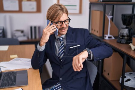 Photo for Young blond man business worker talking on smartphone looking watch at office - Royalty Free Image