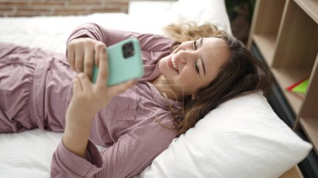 Foto de Joven mujer hispana hermosa usando teléfono inteligente acostado en la cama en el dormitorio - Imagen libre de derechos