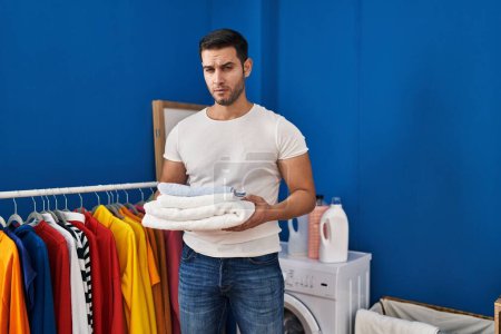 Photo for Young hispanic man with beard holding folded clean towels at laundry room skeptic and nervous, frowning upset because of problem. negative person. - Royalty Free Image