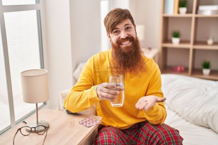 Téléchargez les photos : Young redhead man taking pills drinking water sitting on bed at bedroom - en image libre de droit