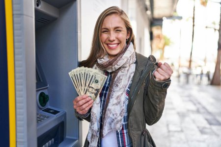 Photo for Young blonde woman holding dollars banknotes from atm machine screaming proud, celebrating victory and success very excited with raised arms - Royalty Free Image