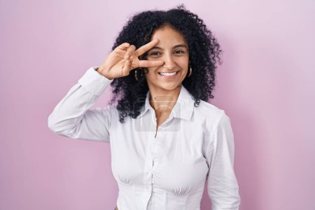 Téléchargez les photos : Hispanic woman with curly hair standing over pink background doing peace symbol with fingers over face, smiling cheerful showing victory - en image libre de droit