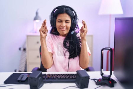 Photo for Mature hispanic woman playing video games at home gesturing finger crossed smiling with hope and eyes closed. luck and superstitious concept. - Royalty Free Image