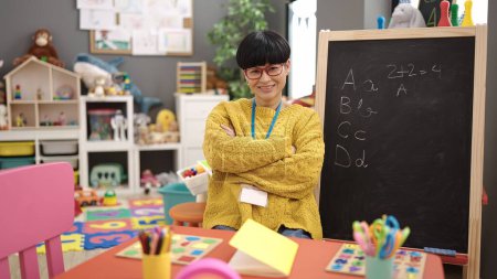 Foto de Young chinese woman preschool teacher smiling confident sitting with arms crossed gesture at kindergarten - Imagen libre de derechos
