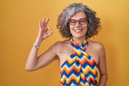 Foto de Mujer de mediana edad con el pelo gris de pie sobre el fondo amarillo sonriendo positiva haciendo signo ok con la mano y los dedos. expresión exitosa. - Imagen libre de derechos